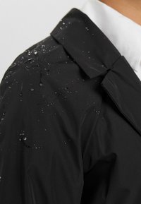 Svart vattentät jacka med en slät textur, med vattendroppar på axelpartiet och en strukturerad krage.