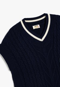 Suéter de punto de color navy con escote en forma de V, presenta acentos de canalé en color crema en el cuello. Patrón de cable texturizado en todo el cuerpo. Etiqueta visible.