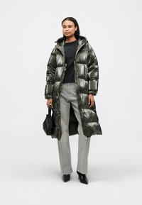 Polstret frakke i mørk olivengrøn, skinnende materiale, knælang med lukning foran, elastiske håndled, parret med grå jeans og sorte accessories.