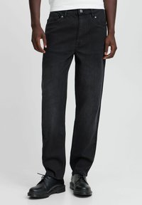 Jeans de mezclilla negra con un diseño de pierna recta, que cuentan con cierre de botón y cremallera, cinco bolsillos y costuras en contraste. Llevados con zapatos negros.