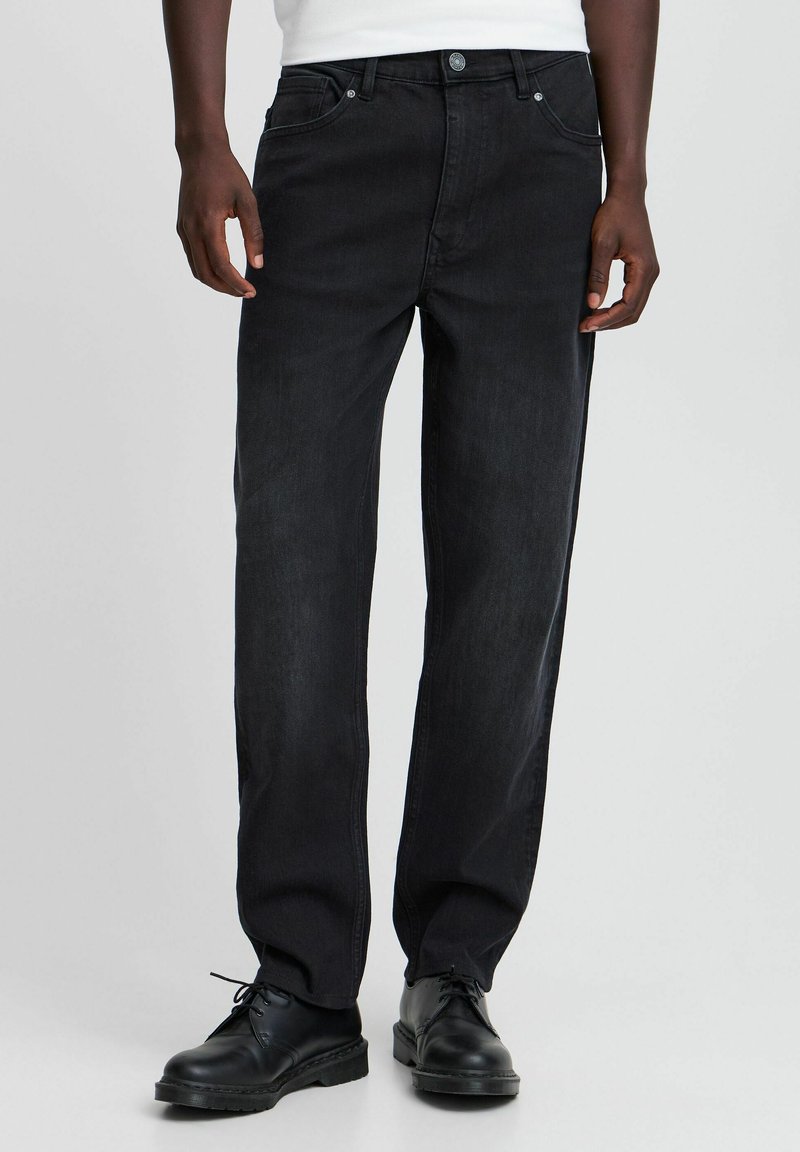Jeans de mezclilla negra con un diseño de pierna recta, que cuentan con cierre de botón y cremallera, cinco bolsillos y costuras en contraste. Llevados con zapatos negros.