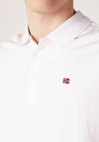 Polo shirt blanc en tissu texturé, doté d'une patte de boutonnage à deux boutons et d'un petit drapeau norvégien brodé sur la poitrine.