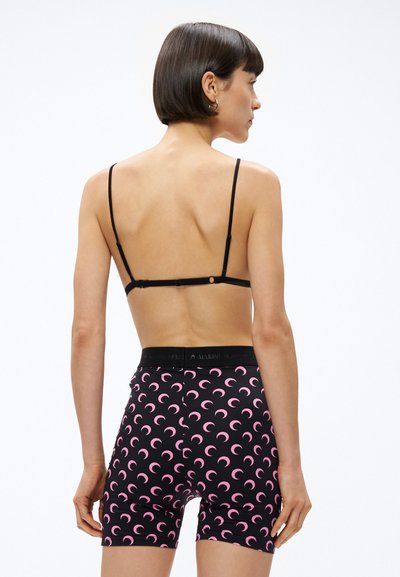 Kobieta z krótkimi ciemnymi włosami, ubrana w czarny biustonosz i czarne szorty z wysokim stanem, ozdobione różowymi wzorami półksiężyców, widziana od tyłu.