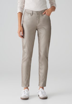 Femme portant un pantalon beige clair slim avec des poches avant, associée à des baskets blanches, debout devant un fond gris uni.