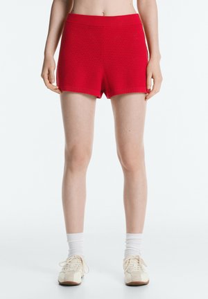 Person iført lyserøde strikkede shorts, hvide tennissokker og beige snøresko står mod en hvid baggrund.