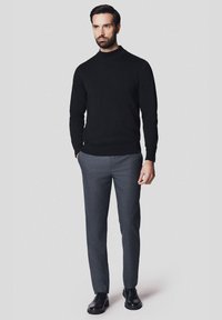 Czarny sweter z dzianiny z wysokim dekoltem, w połączeniu z dopasowanymi szarymi spodniami i czarnymi butami wizytowymi. Model stoi na jednolitym tle.