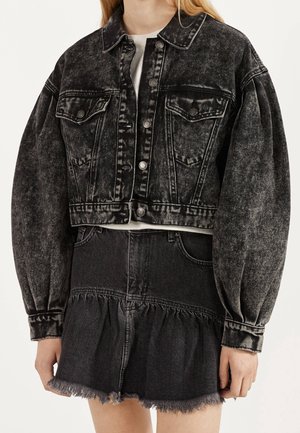 Kobieta w czarnej jeansowej kurtce typu cropped z efektem acid-wash, z bufkami na rękawach oraz dopasowanej, postrzępionej jeansowej spódnicy, na białym topie.