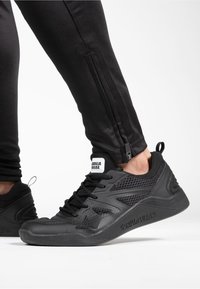 Chaussures de sport noires en matériau mesh et caoutchouc, avec un design texturé, une semelle épaisse et un logo bien visible sur la languette.