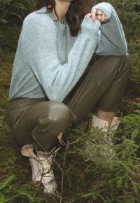 Maglione di lana azzurro, pantaloni in pelle verde oliva e stivaletti bianchi alla caviglia con una fascia, ambientati in uno sfondo naturale con vegetazione.