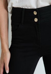 Jeans negros de tiro alto con una textura suave, que presentan dos botones dorados en la parte superior y un bolsillo lateral, diseñados para una silueta ajustada.