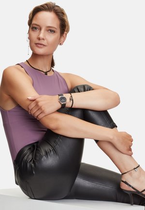 Haut sans manches violet, leggings noirs brillants, montre noire, collier noir en plusieurs couches et boucles d'oreilles en argent ; pose assise avec les jambes croisées.