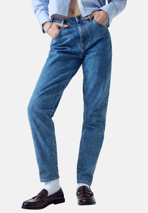 Person trägt hochtaillierte blaue Jeans, hellblaues kurz geschnittenes Hemd, weiße Socken und dunkelbraune Loafer und steht mit den Händen in den Taschen.