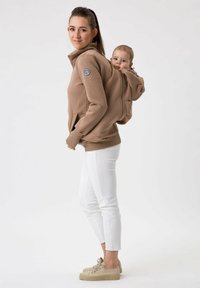 Haut à capuche zippé marron avec une poche arrière pour bébé, doté de poignets et d'un ourlet côtelés. Associé à un pantalon blanc ajusté et des chaussures beiges.