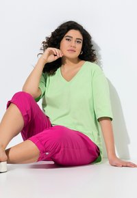 T-shirt verde chiaro a V, a maniche corte, abbinato a pantaloni rosa cropped con dettagli in bottone, seduta su uno sfondo bianco.