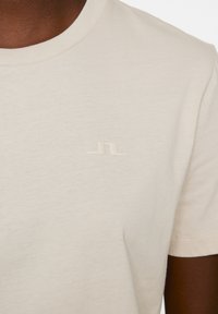 Krämfärgad bomull t-shirt med korta ärmar, med en subtil präglad logotyp på bröstet. Mjuk textur och klassisk rund halsringning.