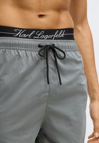 Szürke fürdőnadrág elasztikus derékpánttal, fekete húzózsinórral és Karl Lagerfeld logóval írásszerű betűtípussal. Simogató, könnyű anyag.