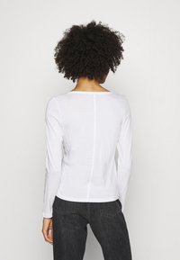 Biała bluzka z długim rękawem o szerokim dekolcie i gładkiej, lekkiej fakturze. Połączona z ciemnymi, dopasowanymi dżinsami z tylnymi kieszeniami.