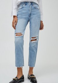 Lichtblauwe high-waisted jeans met gescheurde stukken op de knieën, een rechte pijp en rafelige zomen. Gecombineerd met zwarte instapschoenen.