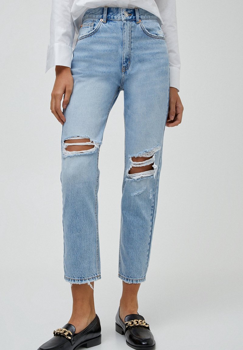 Lichtblauwe high-waisted jeans met gescheurde stukken op de knieën, een rechte pijp en rafelige zomen. Gecombineerd met zwarte instapschoenen.