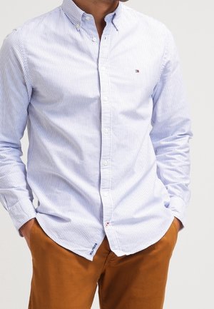 Homme portant une chemise à boutons rayée bleu clair et blanc avec un petit logo sur la poitrine et un pantalon marron, les mains dans les poches.