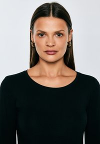 Sirgepruuni juustega naine, kellel on kuldsed rõngakõrvarõngad ja must särk, vaatab otse kaamerasse ühtlase heleda tausta ees.