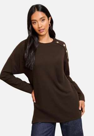 Frau mit langen dunklen Haaren, die einen dunkelbraunen Pullover mit Knöpfen an der Schulter und eine blaue Jeans trägt, posiert mit einer Hand in der Hüfte.