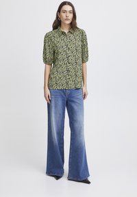 Grünes Blumenmuster-Hemd mit kurzen Puffärmeln, kombiniert mit hellblauen weit geschnittenen Jeans und schwarzen spitz zulaufenden Schuhen.