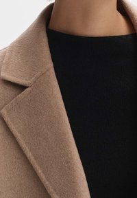 Manteau en laine beige avec une texture douce, présentant un col châle, exposé à côté d'un haut en tricot noir ajusté.