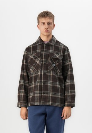 Young man wearing a dark brown plaid button-up jacket with chest pockets and blue pants, standing against a white background.