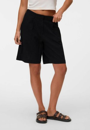 Persona que lleva pantalones cortos negros hasta la rodilla y sandalias oscuras de tiras marrón, ajustando la cintura de los pantalones con una mano.