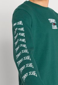 Tummanvihreä pitkähihaiset collegepaita, jossa rinnassa ja vasemmassa hihassa on valkoisilla kirjaimilla "TOMMY JEANS". Valmistettu pehmeästä puuvillamateriaalista.