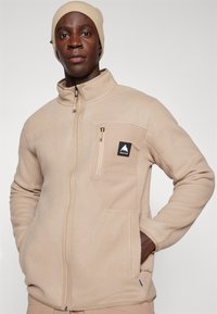 Lys brun fleece-jakke med glidelås foran, høy krage og brystlomme. Har et svart logo-patch og myk tekstur. Varm og funksjonell design.
