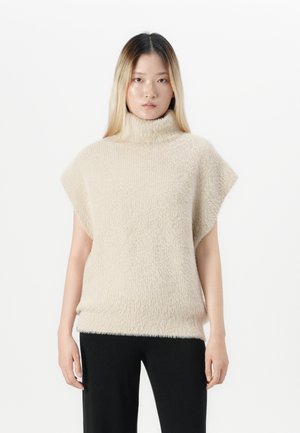 Jonge vrouw in een beige mouwloze coltrui en zwarte broek, staand tegen een effen witte achtergrond.
