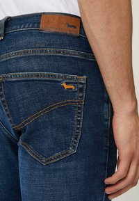 Jeans in denim blu scuro con un'etichetta in pelle marrone con logo in rilievo e cuciture gialle sulla tasca posteriore, che mostra un design di bassotto.