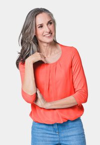 Korallefarbige Bluse mit Dreiviertelärmeln, plissiertem Vorderdesign, weicher Textur und rundem Ausschnitt, kombiniert mit hellblauen Jeans.