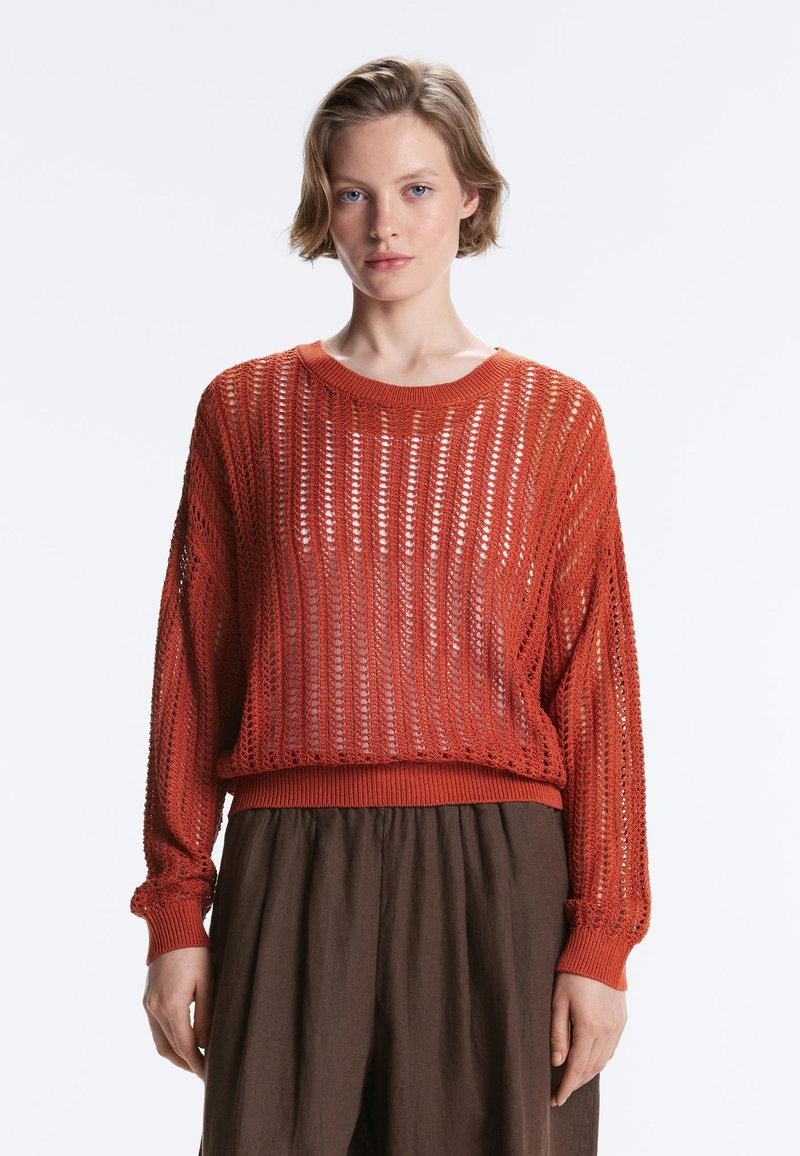 Jeune femme aux cheveux blonds courts portant un pull en maille orange ample sur une jupe marron, debout devant un fond clair uni.