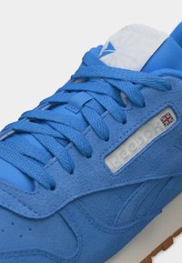 Κοντινό πλάνο μπλε δερμάτινου αθλητικού παπουτσιού με υφασμένες κορδόνια, λευκή ταμπέλα με λογότυπο και μικρή σημαία της Ένωσης Βασιλείων στην πλευρική επιφάνεια.