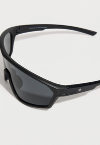 Μαύρα γυαλιά ηλίου με σπορτίβ σχεδιασμό, με σκουρόχρωμους φακούς, ματ φινίρισμα, γωνιώδη πλαίσια και διακριτικό λογότυπο στο σκελετό κοντά στο αυτί.
