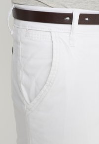 Shorts de mezclilla blanca con una textura suave, que cuentan con bolsillos frontales y un cinturón de cuero marrón. Las costuras delinean los bordes de los bolsillos.