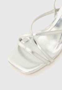 Silberne Metallic-Sandalette mit hohem Absatz, quadratischer Zehenpartie, dünnen überkreuzten Riemen und glatter, reflektierender Oberfläche.