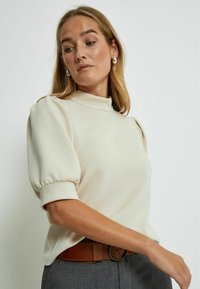 Cream blouse with high neckline and puff sleeves, featuring a smooth texture. Paired with a brown belt and grey trousers.