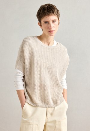 Lys beige strikkegenser med løs passform og korte ermer, lagd over en hvit langermet topp. Strukturert design med ribbestrikket kant.