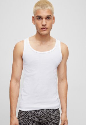 Weißes Baumwoll-Tanktop mit Rundhalsausschnitt, figurbetontem Design und dezentem Branding an der Seite. Kombiniert mit gemusterten grauen Shorts.