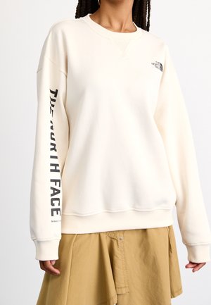 Henkilö, joka käyttää kermaan sävyistä pyöreäkauluksista collegepaitaa, jossa on "The North Face" -logo rinnassa ja hihassa, yhdistettynä ruskean sävyiseen hameeseen.
