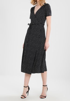 Femme portant une robe portefeuille noire à pois blancs, manches courtes, et des sandales noires à talons hauts avec brides à la cheville, debout sur un fond blanc uni.