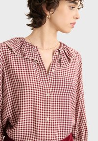 Blusa a quadri rossa e crema con colletto arricciato, patta dei bottoni e maniche lunghe. Il materiale sembra leggero e testurizzato.