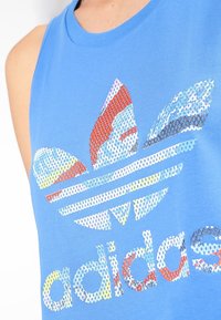 Blå linne med ett stort flerfärgat paljettmönster av Adidas-logotypen med blommiga inslag och horisontella ränder längst ner.