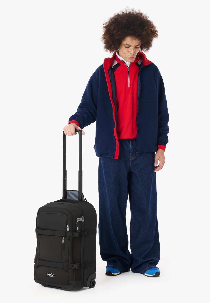 Personne aux cheveux bouclés portant une veste bleue et une chemise rouge, debout en tenant la poignée d'une valise noire à roulettes.