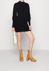 Abito nero a maniche lunghe con collo alto, abbinato a stivaletti in pelle tan con tacco a blocco e zip laterale. Porta una borsa bianca.