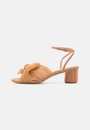 Beige åben tåsandal med plisseret stof, knude foran, ankelrem med spænde og lav blokhæl på hvid baggrund.